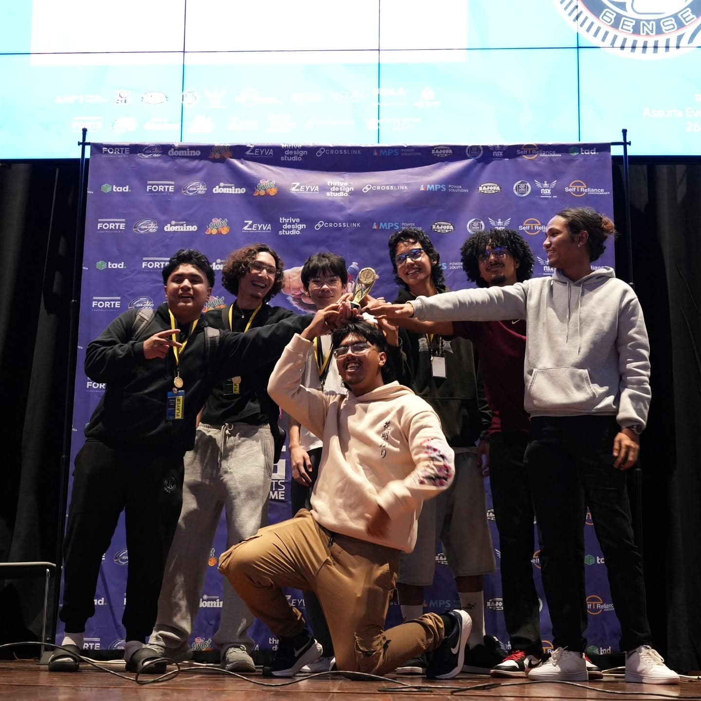The Sixth6ense roster on stage at ESCON 2025, medals round their necks and every hand on the winners' trophy, in front of the event sponsor backdrop